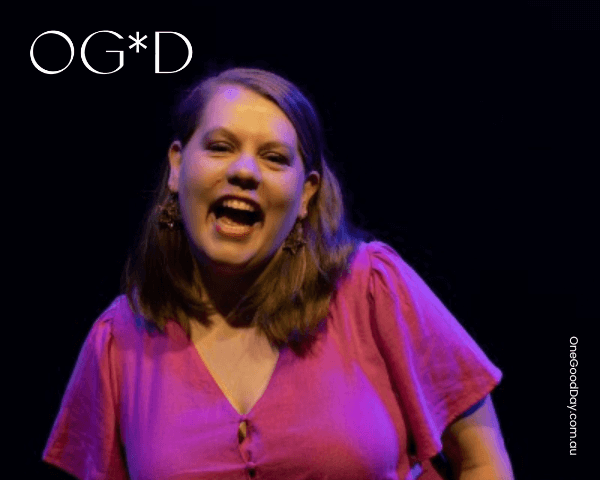 A photo of Alex wearing a magenta top. She is singing or talking on stage with a black background.