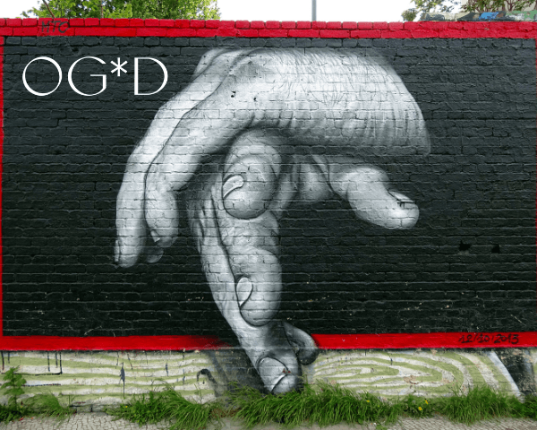 A black brick wall mural featuring a large, grayscale hand gently covering another hand. The top and bottom edges of the wall are painted bright red, creating contrast. Grass grows along the base of the wall. The OG*D logo appears in the bottom left corner.