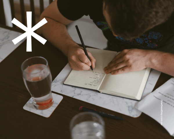 Blog NDIS Psychosocial Disability Evidence A person sits at a table, writing notes in an open notebook. Papers, pens and two glasses of water are arranged around them. A white One Good* Day asterisk appears in the top left corner.