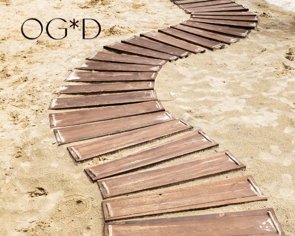 A winding wooden boardwalk curves across pale sand, with the black OG*D logo in the top left corner.